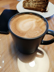 a cup of cappuccino coffee on a table in a cafe next to you can see a plate with a brown cake