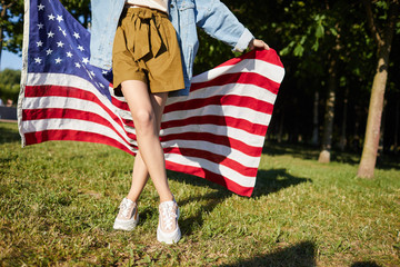 Long legs of unrecognizable woman in beige wide shorts and fashionable sneakers standing on grass in park, woman holding American flag