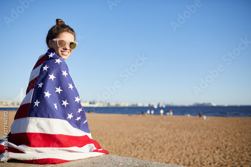 Portrait of happy attractive young woman with hair bun wearing sunglasses covered in big American flag sitting on concrete slab while spending time on beach
