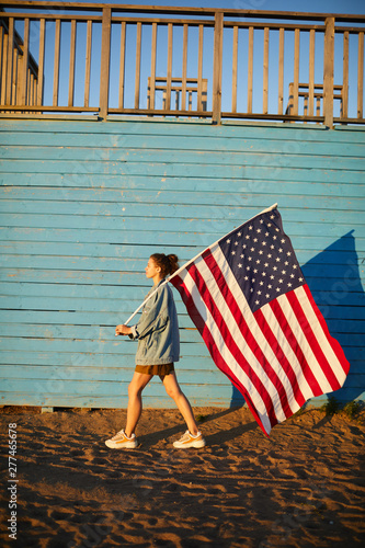 Confident young woman in casual outfit carrying big American flag on stick and going back and forth on beach