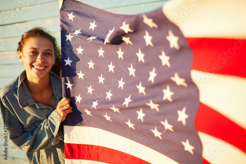 Portrait of happy attractive student girl in denim jacket proud of America holding national flag outdoors