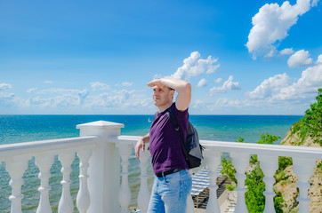 An elderly man stands near a white fence with balusters and looks at the blue sea.