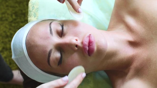 Anti-aging paraffin therapy for face. Facial massage with slices of warm paraffin for young woman in cosmetology spa center. Massagist rubs client's face skin with circular movements, top view.