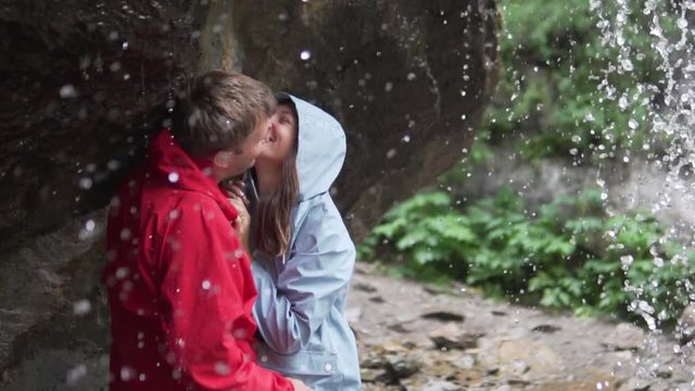 Valentine's Day. Lovers kiss under a waterfall. A couple in a mountain hike stands under a stream of a waterfall in non-wet jackets, they gently kiss and laugh