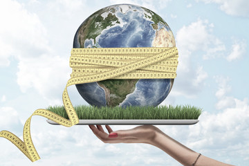 Woman's hand holding digital tablet with grass growing on screen, small Earth on top with yellow...
