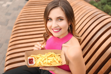 Fototapeta premium Beautiful young woman eating tasty french fries outdoors