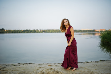 Fototapeta na wymiar Blonde sensual barefoot woman in red marsala dress posing against lake with reeds.
