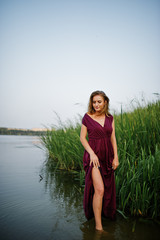 Blonde sensual woman in red marsala dress standing in water of lake with reeds.