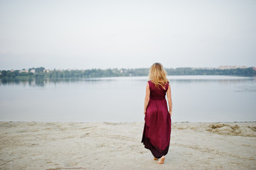 Fototapeta na wymiar Back of blonde sensual barefoot woman in red marsala dress posing against lake.