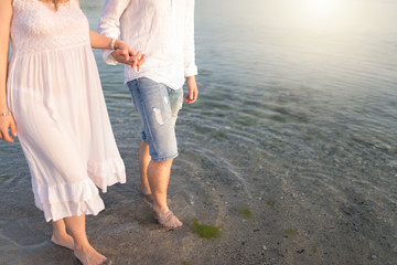 Outdoor shot of romantic young couple walking along the sea shore holding hands. Young man and woman walking on the beach together at sunset, body closeup