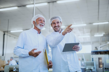 Two smiling Caucasian supervisors in white sterile uniforms standing in food factory. Younger one holding tablet and pointing while older one holding folder with documents under armpit.