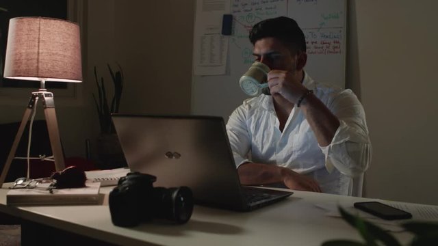 Man Works Back Late in Dark Office at Night, Drinks Coffee at Desk - Wide Shot