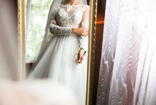 Bride is looking in the vintage mirror. Lace dress with long sleeves and veil. Wedding day details. Elegant morning photo.