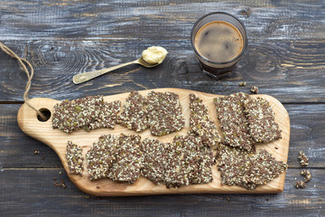 Delicious healthy multigrain gluten-free crackers, ketogenic, from chia seeds, flax, sesame and ground pumpkin seeds on wooden board and keto coffee in glass with spoon of butter оn wooden table
