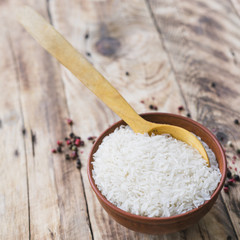 Overhead view of raw white rice bowl with spoon near black pepper spread on wooden table