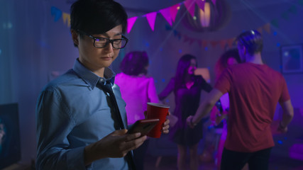 At the Wild House Party: Confident Asian Man Uses Smartphone Instead of Dancing With Other People.