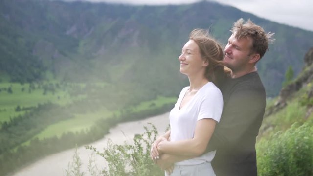 Couple in love on the top of the mountain. A man embraces a woman from behind. They look at the river and the mountains from the peak. Happy family on a mountain hike. Slow shooting