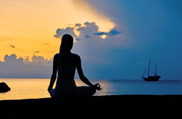  Silhouette of a young woman meditating by the sea against the backdrop of colourful dawn and sail boat