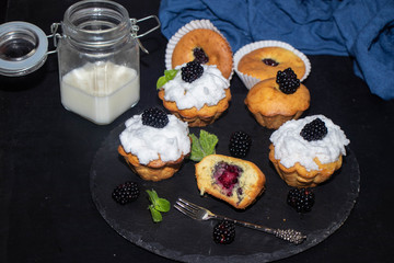 Muffins with white cream and without cream on a black background. Near the bank with milk. Blackberries and mint leaves.