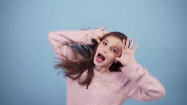 Fun, pretty teen girl in bright sweater, fooling, looking at camera, inflate cheeks, isolated over pastel blue background in studio. People sincere emotions lifestyle concept
