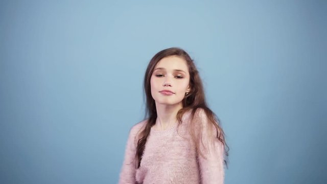 Young pretty long haired girl on a blue background smiling and fooling around and jokingly dancing. Wearing bright pink sweater, looking at the camera