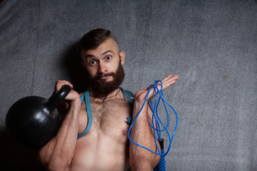 The guy chooses between a jump rope and free weights. Concept cardio or strength. Portrait of a sporty, bearded guy with sport shells, in the studio on a gray background. - obrazy, fototapety, plakaty