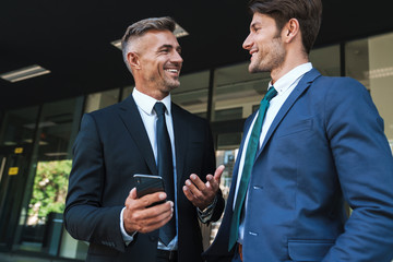 Portrait of happy businessmen partners standing outside job center and using cellphone together...