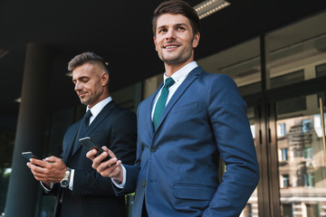 Image of joyful businessmen partners standing outside office center and holding cellphones together during working meeting
