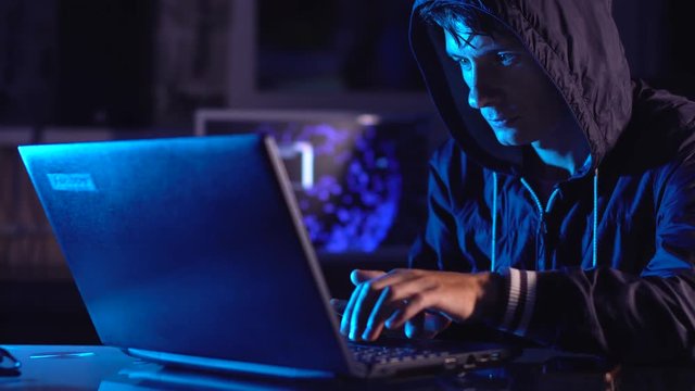 Male hacker in the hood holding the phone in hands trying to hack the mobile device cloud in the dark under neon light