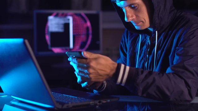 Hacker in the hood holding the phone in his hands trying to hack the mobile device cloud in neon light. Cyber security