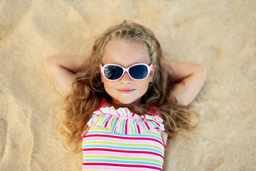Top view of pretty little blonde girl in sunglasses lying on sandy beach during summer vacation