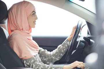 Smiling confident young Muslim lady in pink summer headscarf sitting at steering wheel and looking through windscreen