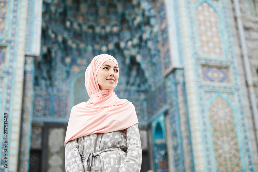 Wall mural Content attractive young Muslim woman in hijab standing against colorful mosque with ornaments and looking away