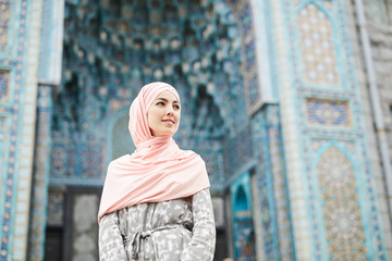 Fototapeta premium Content attractive young Muslim woman in hijab standing against colorful mosque with ornaments and looking away