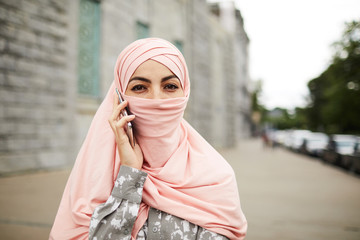 Portrait of successful modern religious Arabian woman in pink hijab standing on street and talking on phone