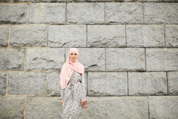 Portrait of content beautiful Muslim lady in summer hijab and coat standing against old building