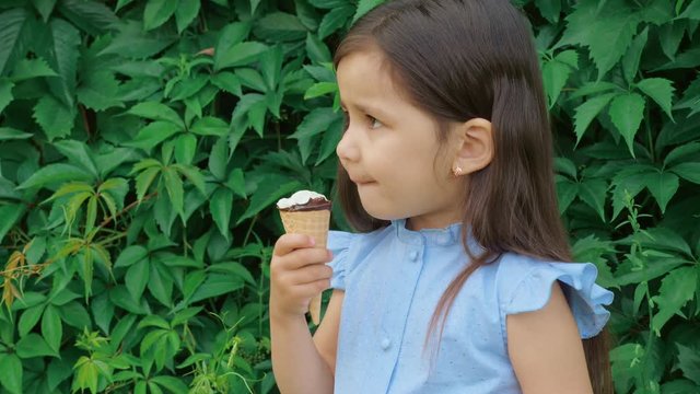 Little cute and funny girl in a blue dress grimaces and eats ice cream. In the background is a green hedge of Virginia creeper (Parthenocissus quinquefolia).