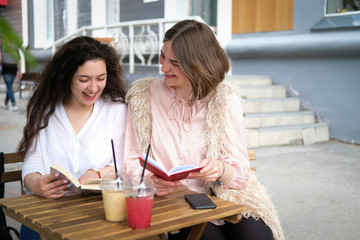 Two young girls reading books at a table in a street cafe. Students preparing for the exam. On the table two summer cocktails in plastic glasses.