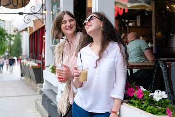 Two young girls are on the street near the summer cafe. Girlfriends communicate and have fun. In the hands of cocktails in plastic glasses.