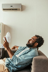 african american man holding blowing electric fan while lying on sofa under air conditioner