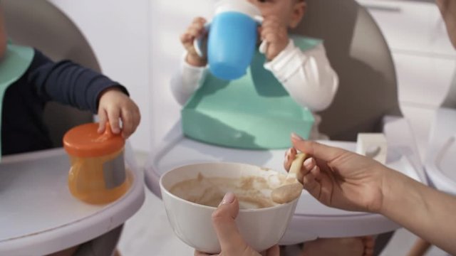Over-shoulder shot of mother mixing baby food in bowl and feeding her 1-year-old mixed race boy, who is drinking from sippy cup, staring away, then opening his mouth and eating, and siblings waiting