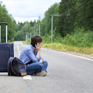 Sad woman with a luggage sits in the middle of a asphalt road and calling on the phone
