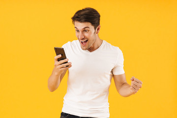 Happy excited shocked young man in casual white t-shirt using mobile phone isolated over yellow background.