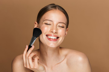 Image of joyful half-naked woman smiling at camera with closed eyes and using makeup brush