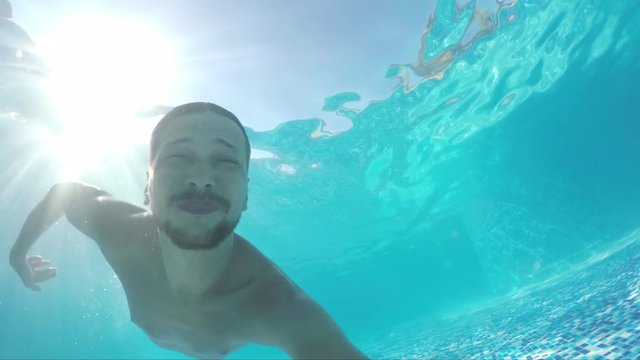 Cute mature bearded slim dark-haired man is swimming under the crystal clear blue water in swimming pool on hot sunny day, view from inside of pool.
