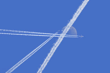 Three contrails and two planes in the blue sky. A strip crosses the moon, which can be seen during the day.