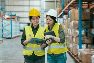 storehouse manager talking with worker in large warehouse. elegant female supervisor teaching new in employee girl while writing on clipboard document. two ladies staff in stockroom wear hard hats.