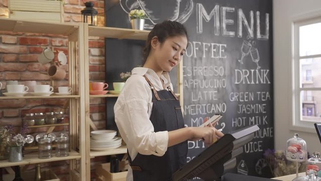 female barista smiling working with tablet behind counter in cafe shop. Coffee culture concepts with real people models. young girl waitress enter customer order in machine hold little note in store.