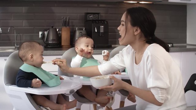 Waist-up shot of middle-aged Filipino mother feeding baby food to her stubborn 1-year-old triplets, who are sitting in high chairs with plastic bibs on necks, unwilling to open mouth, and one crying