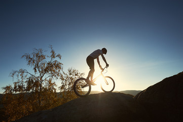 Fototapeta na wymiar Silhouette of male cyclist riding on trial bicycle on top of big boulder, biker making acrobatic stunt on summer evening, blue sky and sunset on background. Concept of extreme sport outdoors